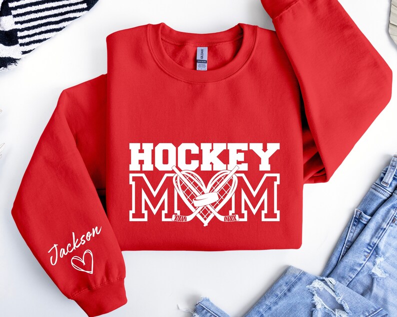 Pu&ograve; includere: Felpa rossa con testo bianco che dice "Hockey Mom" con un bastone da hockey e un disco a forma di cuore. La manica sinistra ha il nome "Jackson" scritto in corsivo con un cuore.