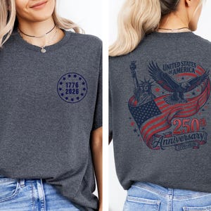 Pode incluir: Camiseta cinza escuro com um pequeno gr&aacute;fico circular azul na frente que diz "1776 2026". A parte de tr&aacute;s da camisa apresenta um design com a Est&aacute;tua da Liberdade, uma &aacute;guia e uma bandeira americana, com o texto "United States of America 250th Anniversary 1776-2026".