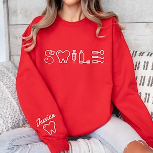 May include: Red sweatshirt with a white graphic of dental tools and the word "Smile" on the front. The sweatshirt has a white tooth graphic on the left sleeve and the name "Jessica" written above it.