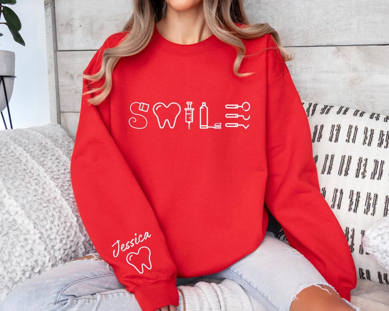 May include: Red sweatshirt with a white graphic of dental tools and the word "Smile" on the front. The sweatshirt has a white tooth graphic on the left sleeve and the name "Jessica" written above it.