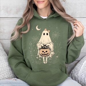 May include: A green hooded sweatshirt with a white ghost holding a jack-o'-lantern. The ghost has a bow on its head and is surrounded by spiderwebs and other Halloween-themed imagery.