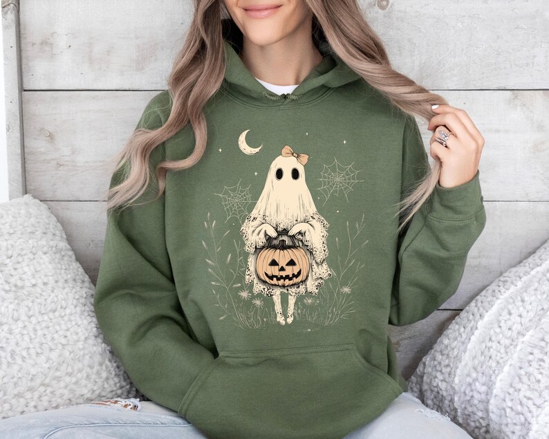 May include: A green hooded sweatshirt with a white ghost holding a jack-o'-lantern. The ghost has a bow on its head and is surrounded by spiderwebs and other Halloween-themed imagery.
