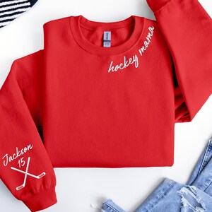 May include: A red crewneck sweatshirt with the words "hockey mama" in white script on the upper right chest. The left sleeve has the name "Jackson" and the number "15" with crossed hockey sticks.