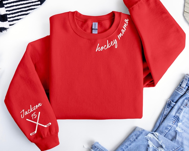 May include: A red crewneck sweatshirt with the words "hockey mama" in white script on the upper right chest. The left sleeve has the name "Jackson" and the number "15" with crossed hockey sticks.
