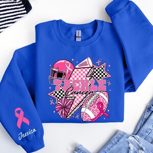 Op de afbeelding: Koningsblauw sweatshirt met een grafisch ontwerp met een roze American footballhelm, voetbal en sterren. De tekst "Tackle Cancer" wordt weergegeven. Een roze lint en de naam "Jessica" zijn ook zichtbaar.