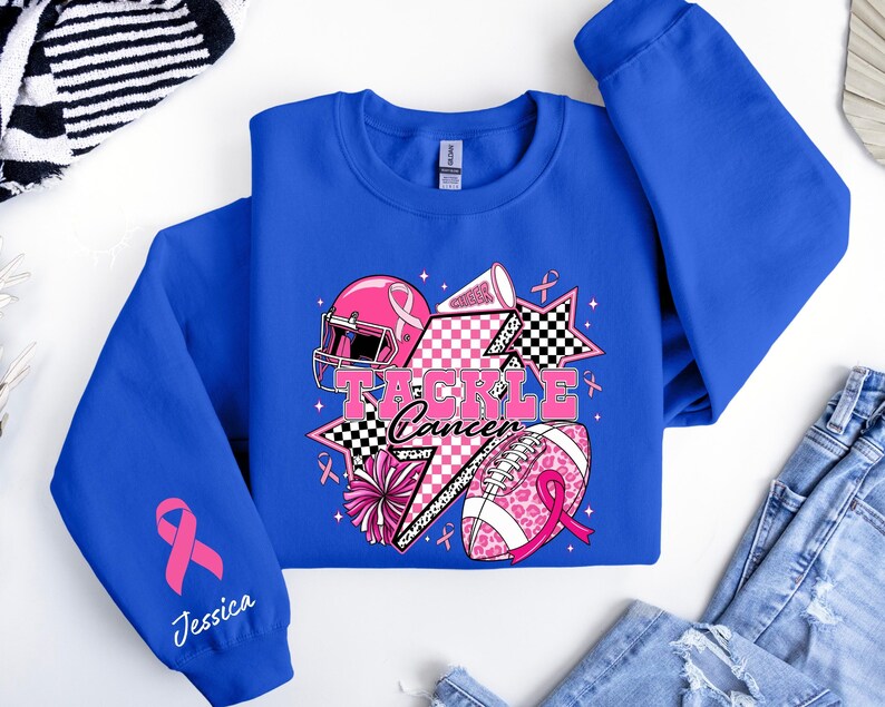 Op de afbeelding: Koningsblauw sweatshirt met een grafisch ontwerp met een roze American footballhelm, voetbal en sterren. De tekst "Tackle Cancer" wordt weergegeven. Een roze lint en de naam "Jessica" zijn ook zichtbaar.