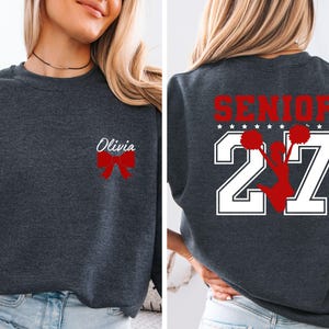 May include: A dark charcoal grey crewneck sweatshirt featuring a red bow and the name Olivia on the front left chest. The back displays the word SENIOR in red above the number 27 in white, with a red silhouette of a cheerleader holding pom-poms.
