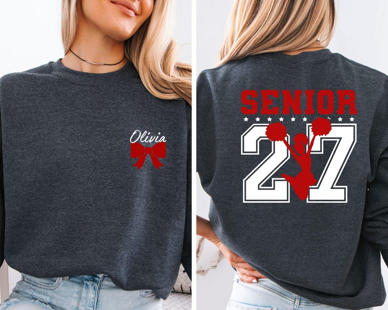 May include: A dark charcoal grey crewneck sweatshirt featuring a red bow and the name Olivia on the front left chest. The back displays the word SENIOR in red above the number 27 in white, with a red silhouette of a cheerleader holding pom-poms.