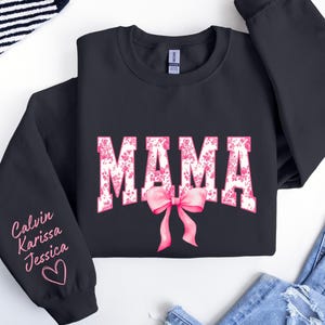 May include: Black sweatshirt with the word "MAMA" in white letters with pink floral accents. A pink bow is below the word. The left sleeve has the names "Calvin, Karissa, Jessica" written in pink with a pink heart.