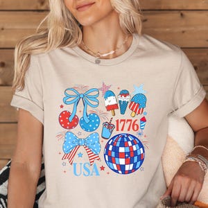 Puede incluir: Camiseta beige con un dise&ntilde;o gr&aacute;fico colorido. El dise&ntilde;o incluye elementos patri&oacute;ticos como lazos, helados, una bola de discoteca y los textos "USA" y "1776" en rojo, blanco y azul.