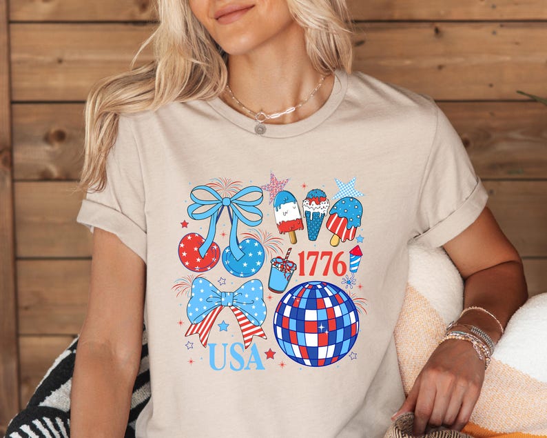Puede incluir: Camiseta beige con un dise&ntilde;o gr&aacute;fico colorido. El dise&ntilde;o incluye elementos patri&oacute;ticos como lazos, helados, una bola de discoteca y los textos "USA" y "1776" en rojo, blanco y azul.