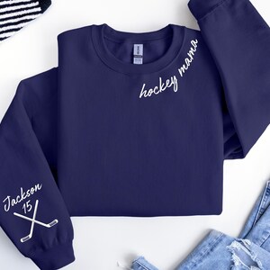 May include: Navy blue sweatshirt with white text reading "hockey mama" and "Jackson 15" with crossed hockey sticks. The sweatshirt is a crew neck style and has long sleeves. The sleeves have the text and graphic on them.