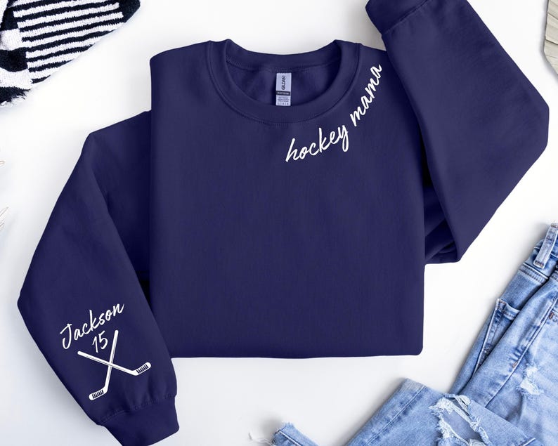 May include: Navy blue sweatshirt with white text reading "hockey mama" and "Jackson 15" with crossed hockey sticks. The sweatshirt is a crew neck style and has long sleeves. The sleeves have the text and graphic on them.
