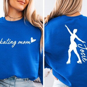 May include: A royal blue sweatshirt with the text "skating mom" and a small heart on the front. The back of the sweatshirt features a white silhouette of a figure skater with the name "Josie" written next to it.