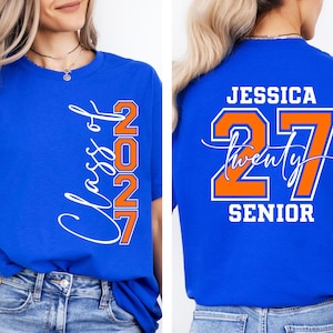 May include: Royal blue t-shirt with the text "Class of 2027" in white script and orange block letters. The back of the shirt features the name "Jessica", the number "27", and the word "Senior" in white and orange.