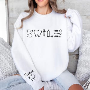 May include: White sweatshirt with a black graphic of a tooth, syringe, toothbrush, and floss with the word "Smile" written in a stylized font. The sweatshirt has the name "Jessica" written on the sleeve with a small tooth graphic.