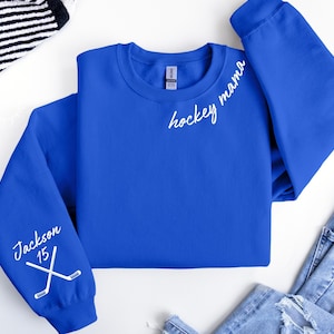 May include: A royal blue sweatshirt with the words "hockey mama" in white script on the chest. The left sleeve features the name "Jackson" and the number "15" with crossed hockey sticks. The sweatshirt is folded on a white surface.