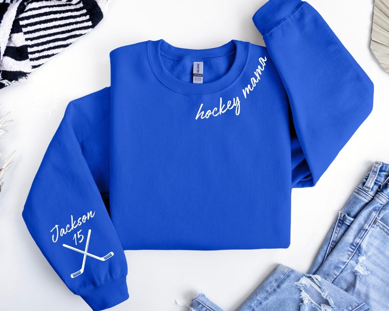 May include: A royal blue sweatshirt with the words "hockey mama" in white script on the chest. The left sleeve features the name "Jackson" and the number "15" with crossed hockey sticks. The sweatshirt is folded on a white surface.