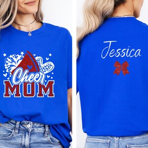 Puede incluir: Camiseta azul con un dise&ntilde;o de porrista rojo, blanco y azul que dice "Cheer Mom". La parte posterior de la camiseta tiene un lazo rojo y el nombre "Jessica" escrito en blanco.