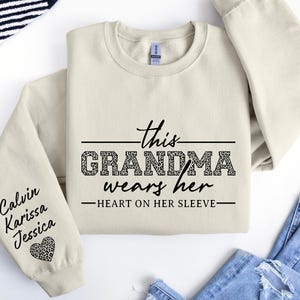 May include: A cream-colored sweatshirt with the text "This Grandma wears her heart on her sleeve" in black. The sleeve has the names "Calvin, Karissa, Jessica" written in black with a leopard print heart.