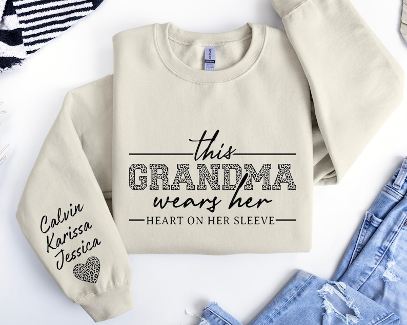 May include: A cream-colored sweatshirt with the text "This Grandma wears her heart on her sleeve" in black. The sleeve has the names "Calvin, Karissa, Jessica" written in black with a leopard print heart.