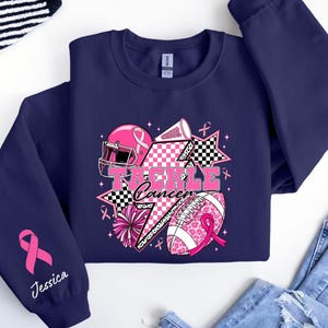 Op de afbeelding: Marineblauw sweatshirt met een roze en zwarte afbeelding. De afbeelding bevat een footballhelm, een football en de woorden "Tackle Cancer". Een roze lint en de naam "Jessica" staan op de mouw.