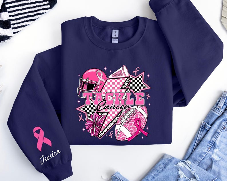 Op de afbeelding: Marineblauw sweatshirt met een roze en zwarte afbeelding. De afbeelding bevat een footballhelm, een football en de woorden "Tackle Cancer". Een roze lint en de naam "Jessica" staan op de mouw.