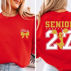 May include: A red crewneck sweatshirt featuring a gold bow and the name Olivia on the front left chest. The back displays the word SENIOR in gold, the number 27 in large white font with gold outlines, and a gold silhouette of a cheerleader.