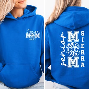 May include: A royal blue hoodie with white text and a front pocket. The front of the hoodie says "Senior Mom 2027" with a cheerleader graphic. The back of the hoodie says "Senior Mom Sierra 2027" with a cheerleader graphic.