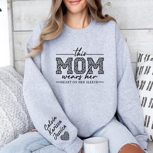 May include: Gray sweatshirt with the text "This Mom wears her heart on her sleeve" in black. The word "Mom" is decorated with a leopard print pattern. The sleeves have the names "Calvin, Karissa, Jessica" written in black.