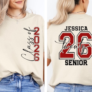 May include: Cream-colored t-shirt with "Class of 2026" in black script and red block letters. The back of the shirt features the name "Jessica", the number "26", and the word "Senior" in black and red.