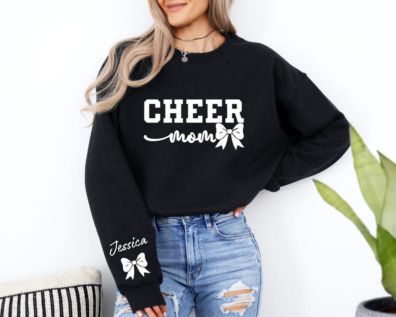 May include: Black sweatshirt with the text "Cheer Mom" and a white bow. The sweatshirt has a white bow and the name "Jessica" on the sleeve.
