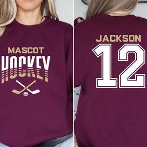 K&ouml;nnte beinhalten: Burgunderfarbener Sweatshirt mit wei&szlig;em Text, der "Mascot Hockey" lautet, und einer goldenen Grafik eines Hockeyschl&auml;gers. Die R&uuml;ckseite des Sweatshirts hat wei&szlig;en Text, der "Jackson 12" in einer gro&szlig;en, fetten Schrift lautet.