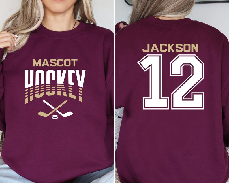 K&ouml;nnte beinhalten: Burgunderfarbener Sweatshirt mit wei&szlig;em Text, der "Mascot Hockey" lautet, und einer goldenen Grafik eines Hockeyschl&auml;gers. Die R&uuml;ckseite des Sweatshirts hat wei&szlig;en Text, der "Jackson 12" in einer gro&szlig;en, fetten Schrift lautet.