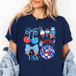 Puede incluir: Camiseta azul marino con un dise&ntilde;o patri&oacute;tico con lazos, estrellas y helados en rojo, blanco y azul. Tambi&eacute;n se incluye la palabra "USA" y el n&uacute;mero "1776".