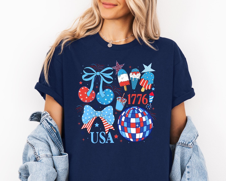Puede incluir: Camiseta azul marino con un dise&ntilde;o patri&oacute;tico con lazos, estrellas y helados en rojo, blanco y azul. Tambi&eacute;n se incluye la palabra "USA" y el n&uacute;mero "1776".