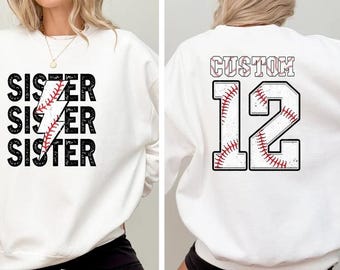 May include: White sweatshirt with the word "Sister" repeated three times with a lightning bolt made of a baseball graphic. The back of the sweatshirt has a white baseball with red stitching and the number "12" with the word "Custom" above it.