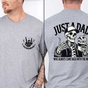 May include: A light gray t-shirt featuring a skeleton graphic. The front of the shirt has a small design with the word "FATHERHOOD". The back of the shirt has a skeleton wearing a hat and holding a milk carton with the text "JUST A DAD WHO ALWAYS CAME BACK WITH THE MILK."