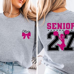 May include: A heather grey crewneck sweatshirt featuring a small pink bow graphic and the name Olivia on the front left chest. The back displays the word SENIOR in pink above the number 27 in black with a pink cheerleader silhouette in the centre.