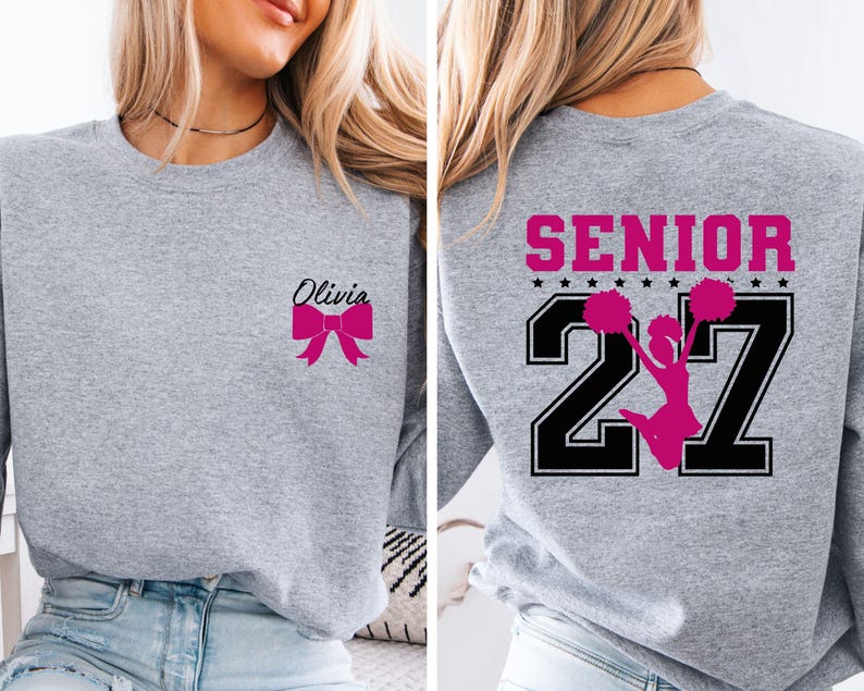 May include: A heather grey crewneck sweatshirt featuring a small pink bow graphic and the name Olivia on the front left chest. The back displays the word SENIOR in pink above the number 27 in black with a pink cheerleader silhouette in the centre.