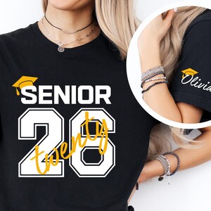 May include: Black t-shirt with "SENIOR 20" in white and yellow lettering, featuring a graduation cap graphic. The sleeve has a graduation cap graphic and the name "Olivia" in white.