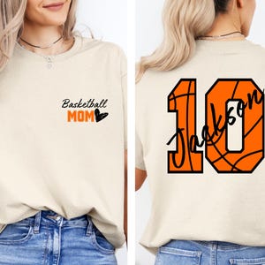 Peut inclure: Un t-shirt beige avec le texte "Basketball Mom" et un c&oelig;ur sur le devant. Le dos du t-shirt a un num&eacute;ro 10 sur le th&egrave;me du basketball avec le nom "Jackson" &eacute;crit &agrave; l'int&eacute;rieur du ballon de basket.