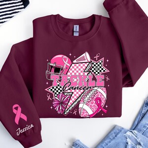 Op de afbeelding: Bordeaux sweatshirt met roze voetbalthema-elementen en de tekst "Tackle Cancer". Het ontwerp bevat een roze helm, een voetbal en cheerleading-elementen. Een roze lint en de naam "Jessica" staan op de mouw.