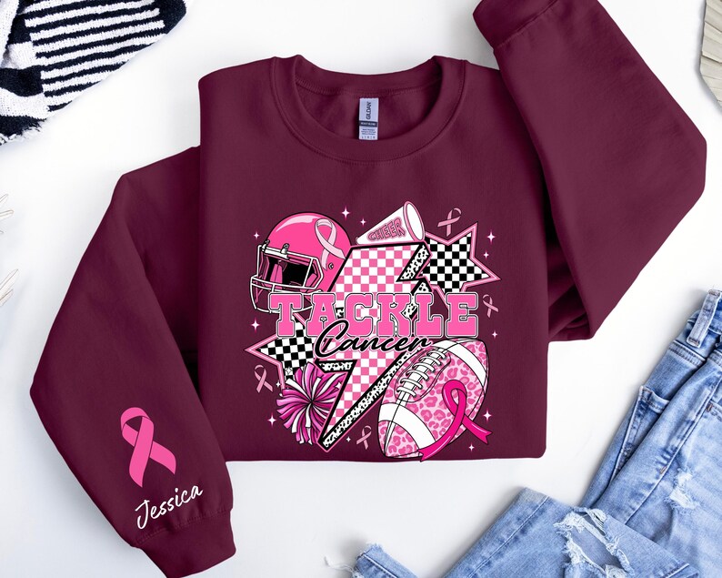 Op de afbeelding: Bordeaux sweatshirt met roze voetbalthema-elementen en de tekst "Tackle Cancer". Het ontwerp bevat een roze helm, een voetbal en cheerleading-elementen. Een roze lint en de naam "Jessica" staan op de mouw.
