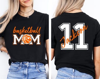 Puede incluir: Camiseta negra con "basketball MOM" en letras naranjas y blancas, con un gráfico de baloncesto. La parte trasera de la camiseta muestra el número "11" en letras blancas con "Jackson" en naranja.