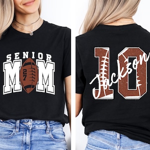 May include: A black t-shirt featuring a front graphic with the text SENIOR MOM and a brown football with the number 27. The back graphic displays the number 10 with a football texture and the name Jackson written in white script across the center.