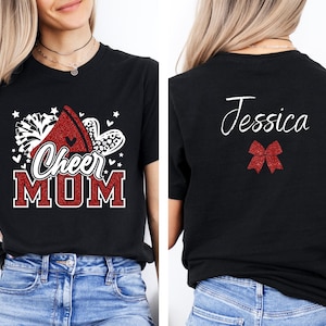 Puede incluir: Camiseta negra con texto rojo de purpurina que dice "Cheer Mom" y un pomp&oacute;n rojo y blanco de purpurina. La parte posterior de la camiseta tiene el nombre "Jessica" en purpurina blanca y un lazo rojo de purpurina.