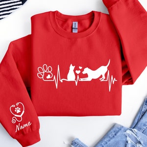 May include: Red sweatshirt with a white graphic of a cat and dog with a heartbeat line and paw print. The left sleeve has a stethoscope and paw print graphic with the word "Name" underneath.