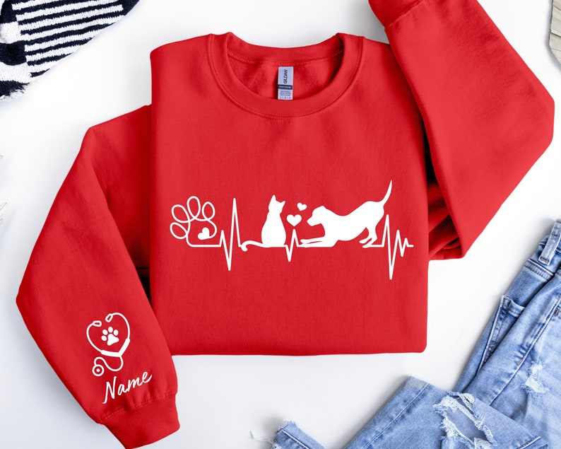 May include: Red sweatshirt with a white graphic of a cat and dog with a heartbeat line and paw print. The left sleeve has a stethoscope and paw print graphic with the word "Name" underneath.