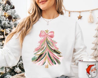 Könnte beinhalten: Weißer Pullover mit einem pinkfarbenen und grünen Weihnachtsbaumdesign. Der Baum hat eine große rosa Schleife und goldene Glitzerakzente.